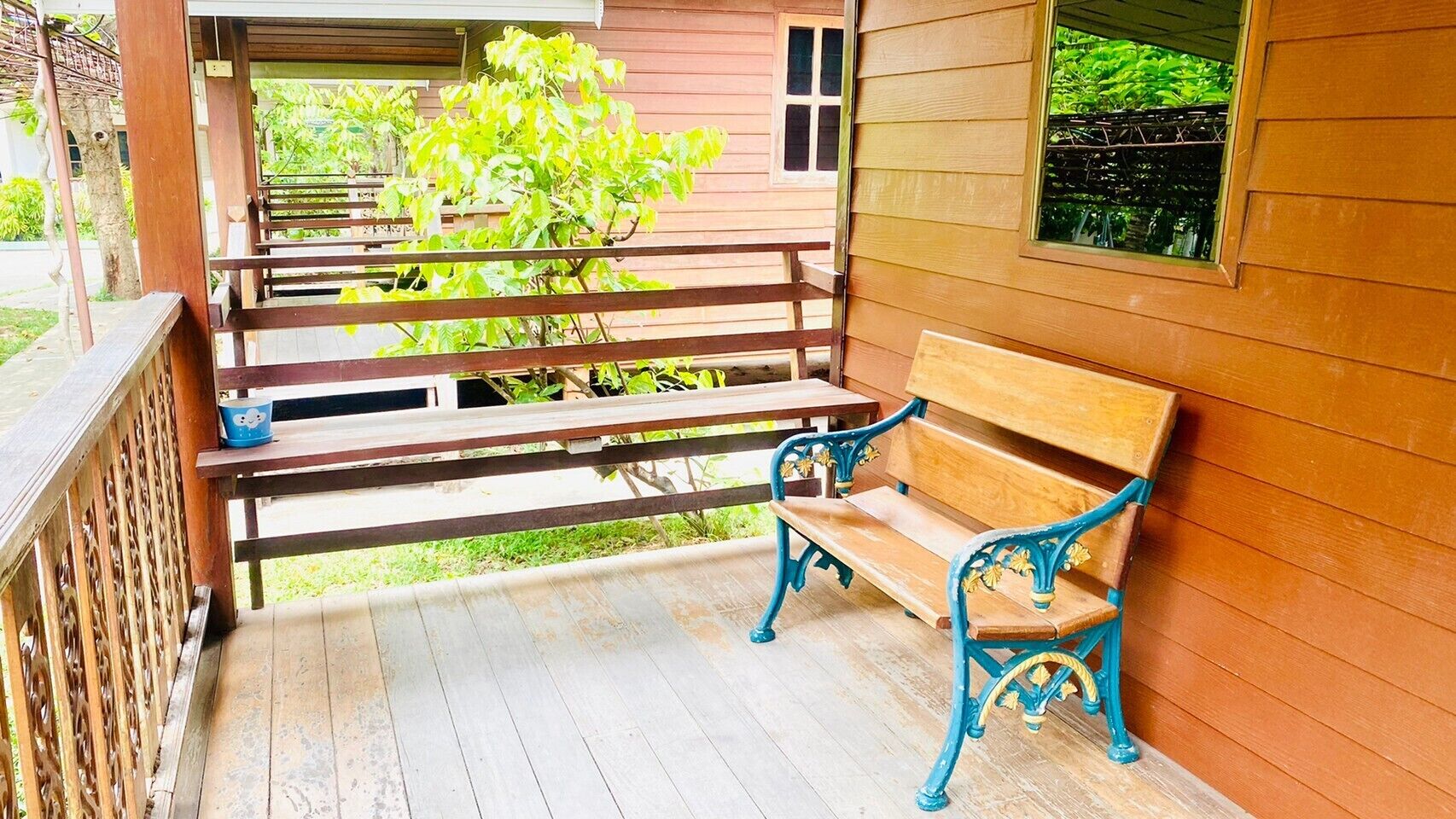 A wooden porch with a bench and a table, overlooking a garden and a building.