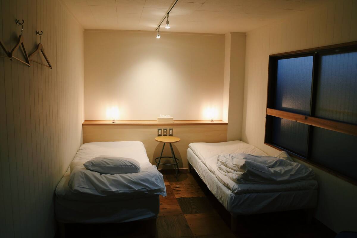 Ein kleiner, schummrig beleuchteter Raum mit zwei Einzelbetten, einem kleinen Tisch und einem Fenster mit Jalousien.
