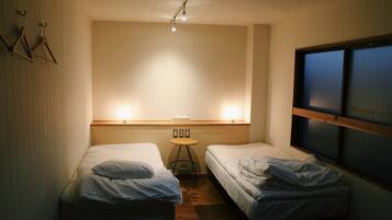 A small, dimly lit room with two single beds, a small table, and a window with blinds.