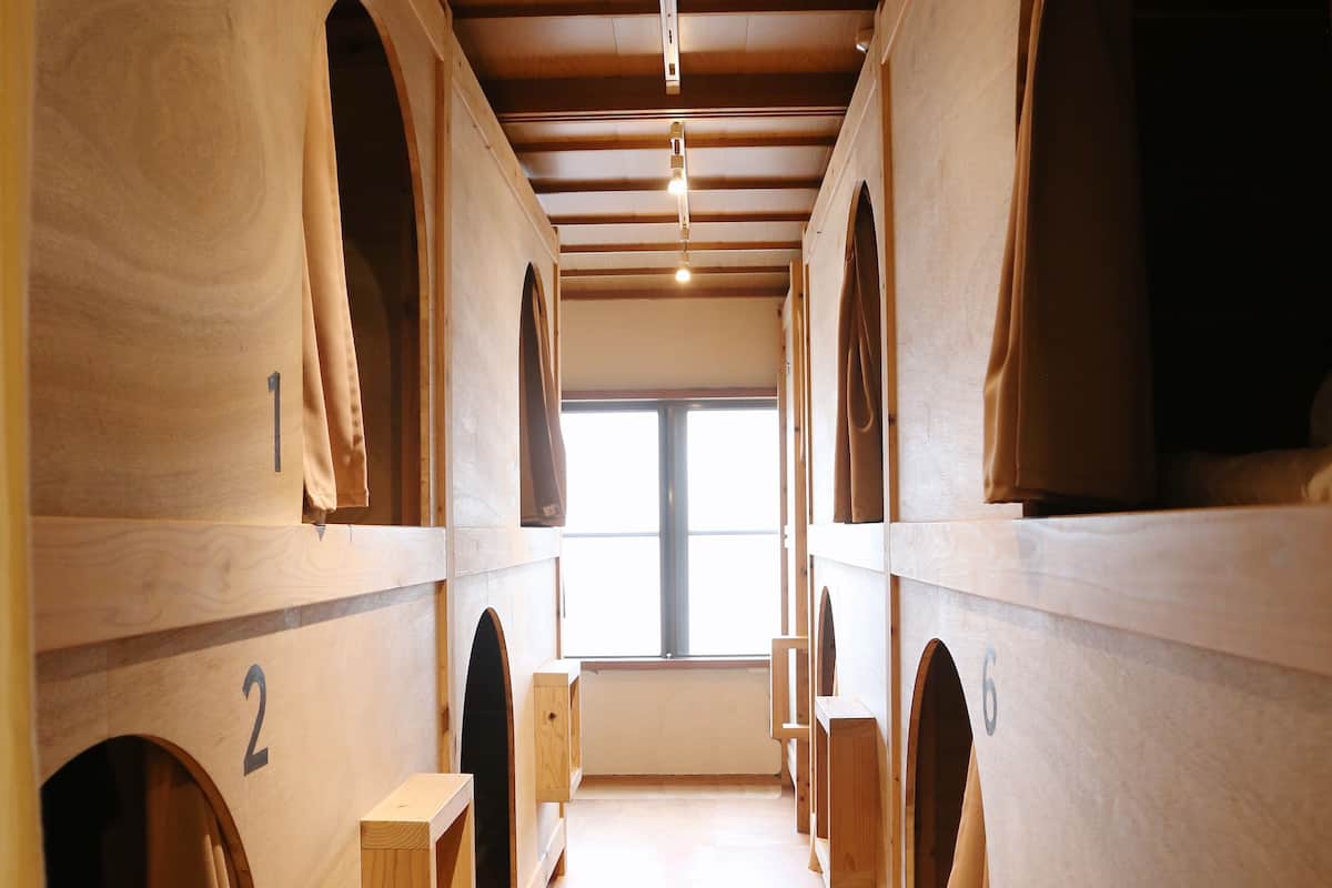 Reihenweise hölzerne Etagenbetten mit den Nummern 1 bis 6, ein Holzboden und ein Fenster am Ende des Raumes.