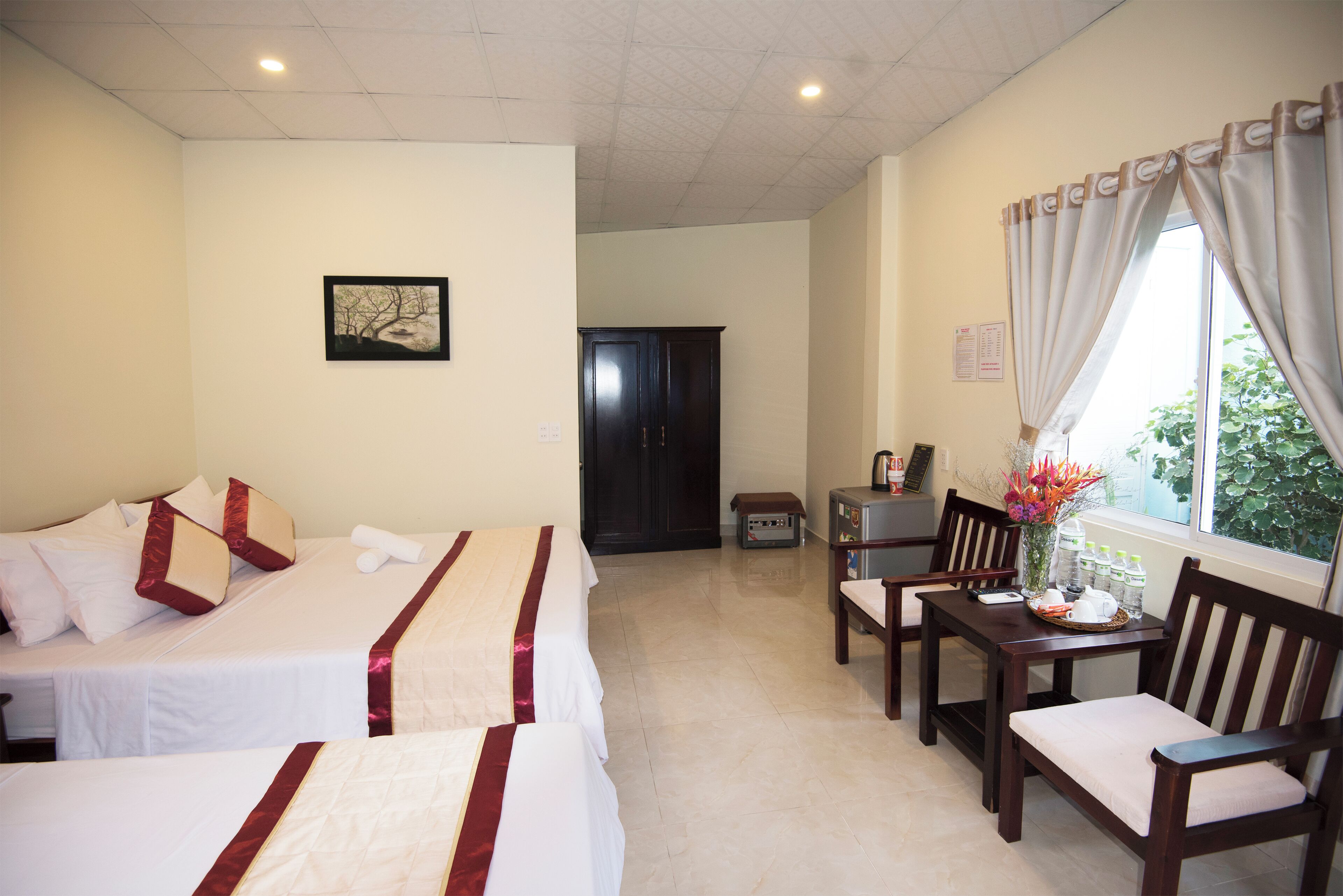 A hotel room with two beds, a dining table with chairs, a small cabinet, and a window with curtains.