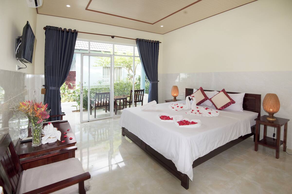 A spacious bedroom with a large bed, a bedside table with a lamp, a TV mounted on the wall, and a view of the outdoors through a sliding glass door.