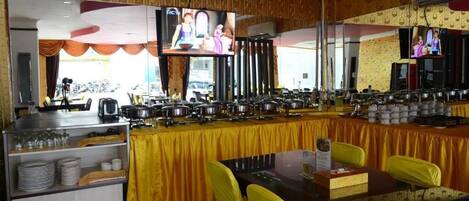 A buffet setup with multiple serving stations, a television mounted on the wall, and yellow chairs around a dining table.