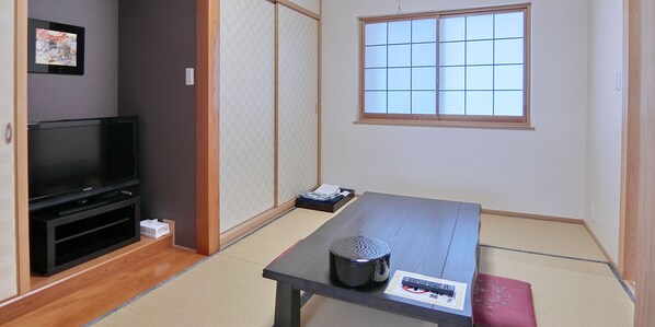 Une pièce de style japonais traditionnel, avec un sol recouvert de tatamis, une petite table avec une télécommande, un téléviseur posé sur un meuble et une porte coulissante.