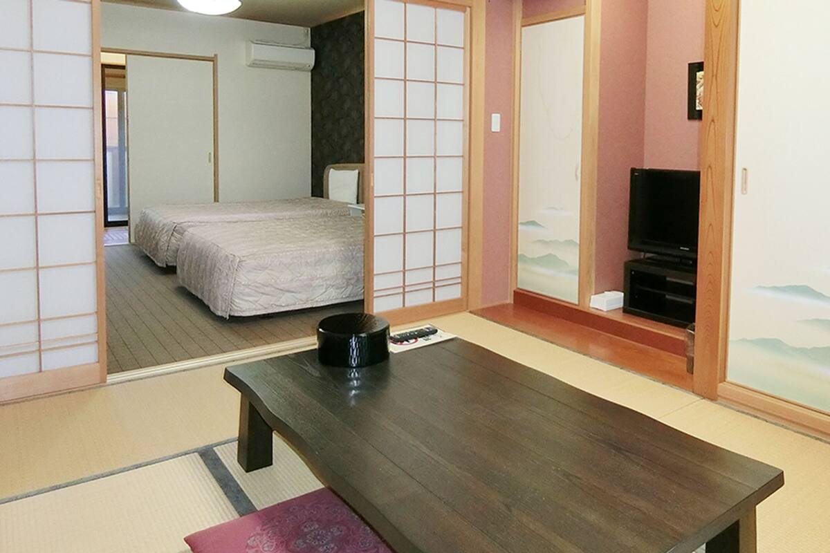 A traditional Japanese-style room with tatami flooring, a low wooden table, a bed with a futon, and a television on a low stand.