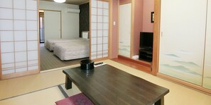 Una habitación tradicional japonesa con suelo de tatami, una mesa de madera baja, una cama con futón y un televisor sobre un mueble bajo.