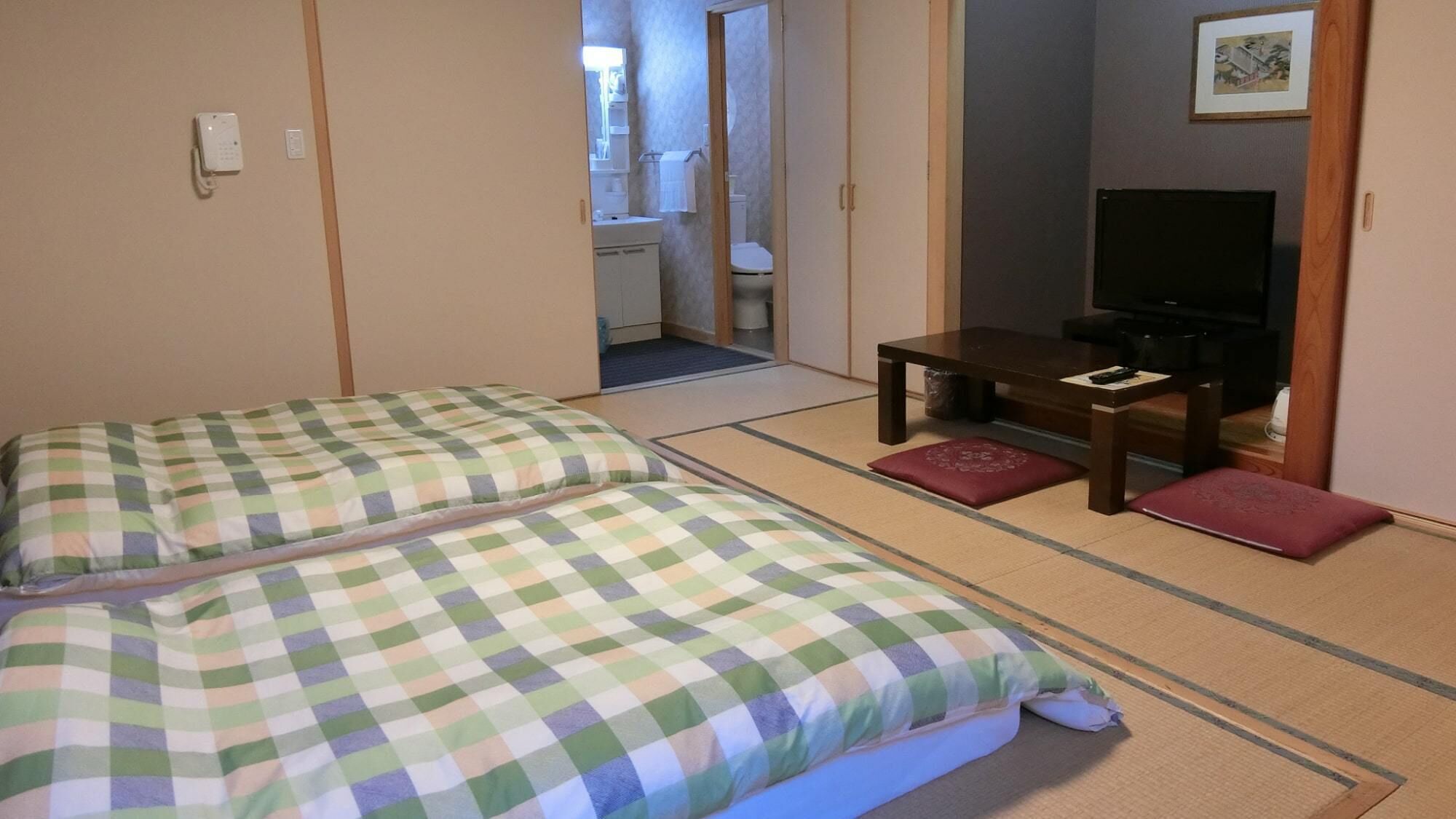 A traditional Japanese-style room with tatami flooring, a bed with checkered bedding, a small table with a TV, and a bathroom visible through an open door.