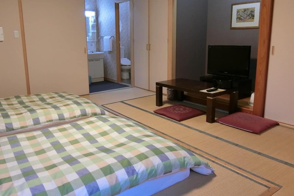 Een traditionele Japanse kamer met tatami vloerbedekking, een bed met geruit beddengoed, een kleine tafel met een televisie, en een badkamer zichtbaar door een geopende deur.