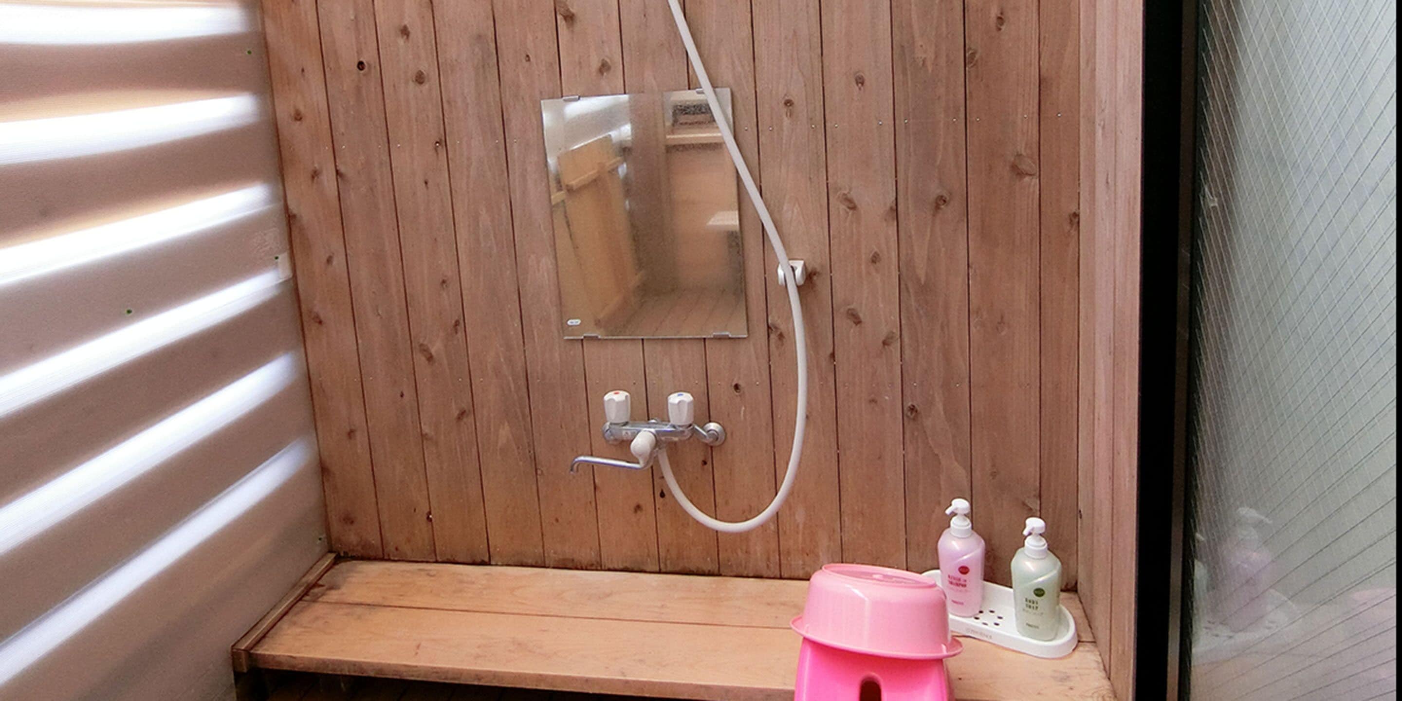 A wooden shower enclosure with a bench, a glass shower door, and two bottles of soap on a shelf.