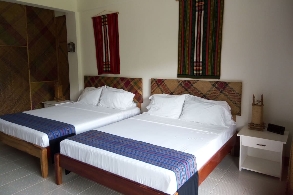 Habitación de hotel con dos camas, cada una con sábanas blancas y una colcha azul a rayas. Hay una mesita de noche de madera con una lámpara pequeña y un control remoto. La habitación tiene un ventanal grande con una cortina colorida y un cabecero con estampado de cuadros.