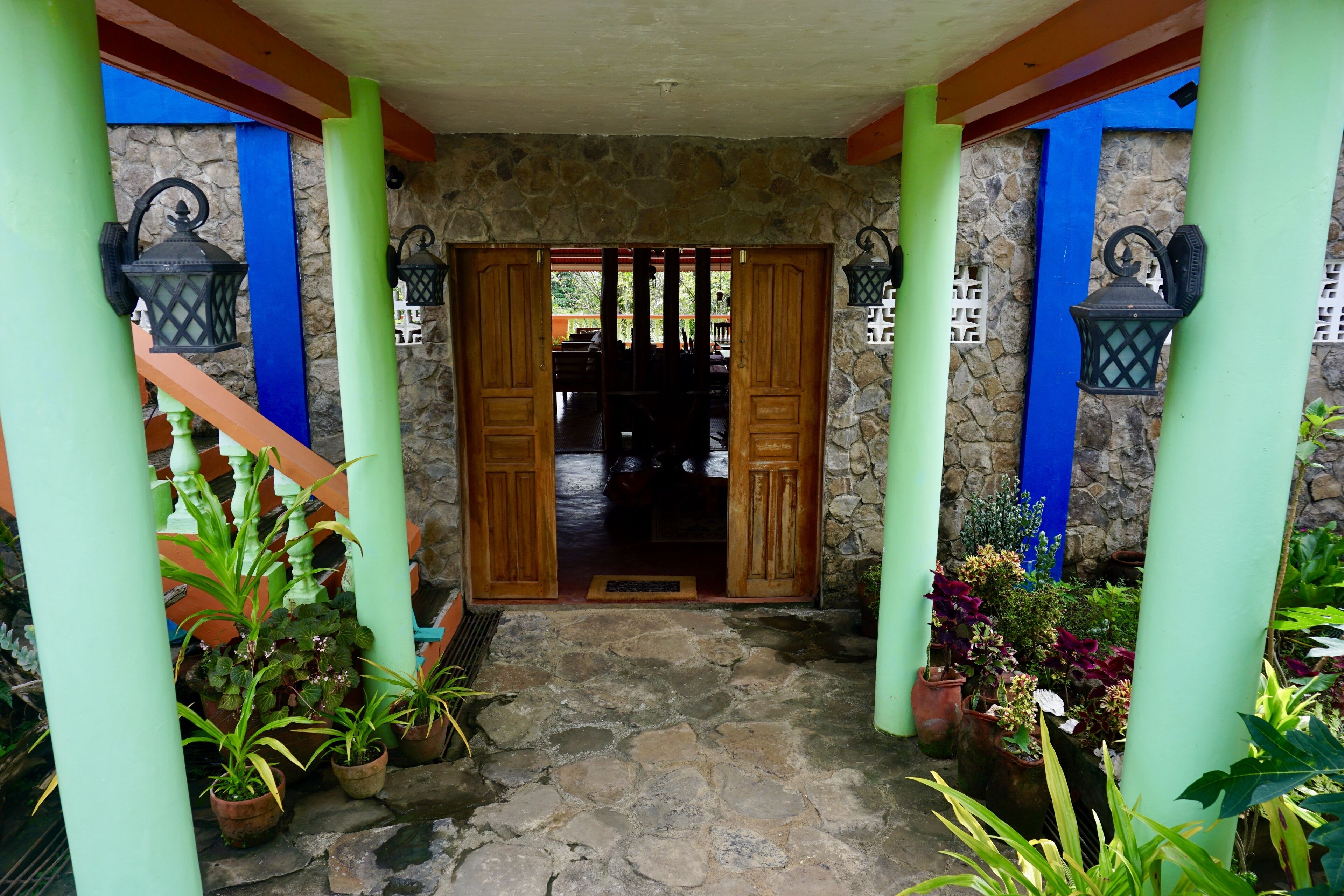 A stone-floored entrance with wooden doors and green pillars, flanked by potted plants and outdoor lighting fixtures.