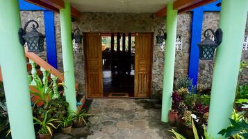 A stone-floored entrance with wooden doors and green pillars, flanked by potted plants and outdoor lighting fixtures.