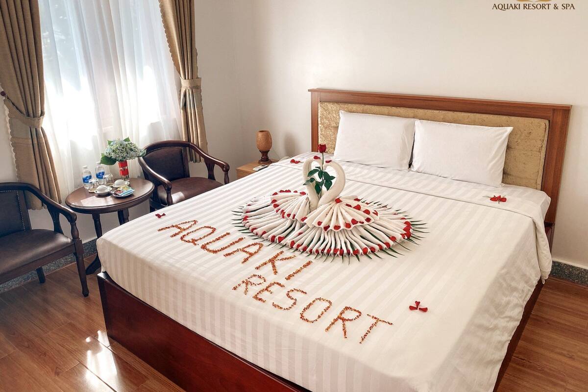 A hotel room with a bed, a wooden headboard, a nightstand, a chair, a small table with a vase of flowers, and a window with curtains.