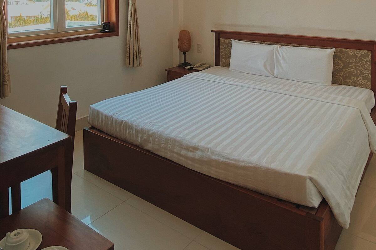 A hotel room with a large bed, a desk with a chair, a window with curtains, and an air conditioning unit.
