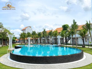 A circular swimming pool with a stone border, surrounded by palm trees and a white building with a red roof. - Aquaki Resort & Spa (Ha Tien)