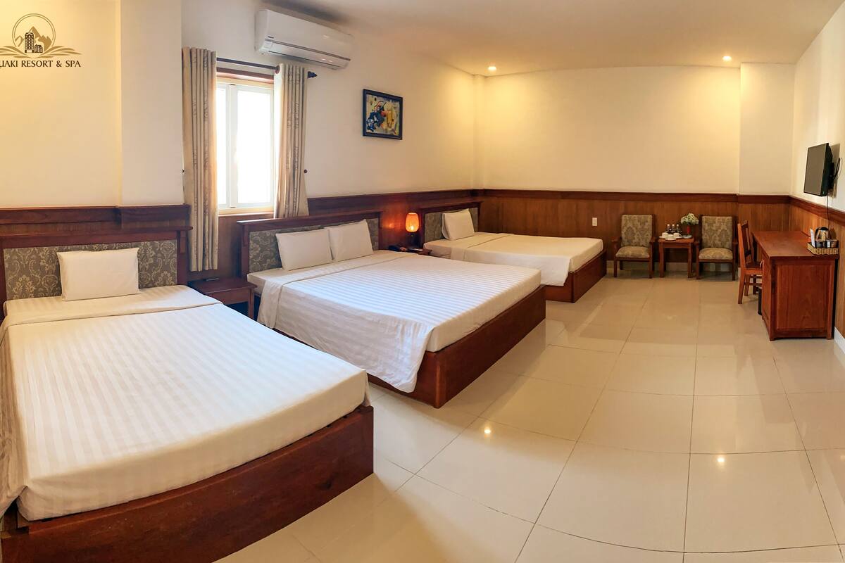 A hotel room with three beds, a wooden headboard, a small table with chairs, a desk, and a television.