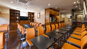 A restaurant with long tables and orange chairs, a kitchen area, and a TV screen. - Hotel Sealuck Pal Kofu (Kofu)