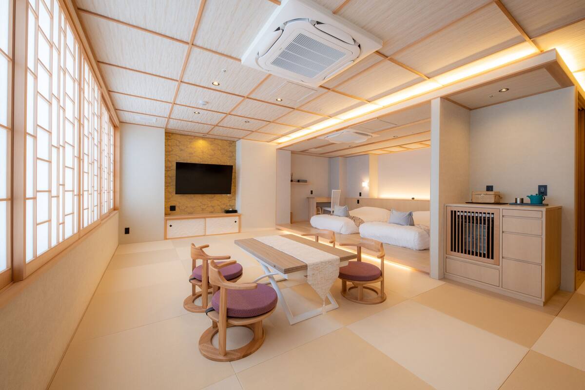 Una habitación moderna y bien iluminada, con techo de madera, televisor de pantalla plana en la pared, un conjunto de sofás y una pequeña zona de comedor con mesa y sillas.