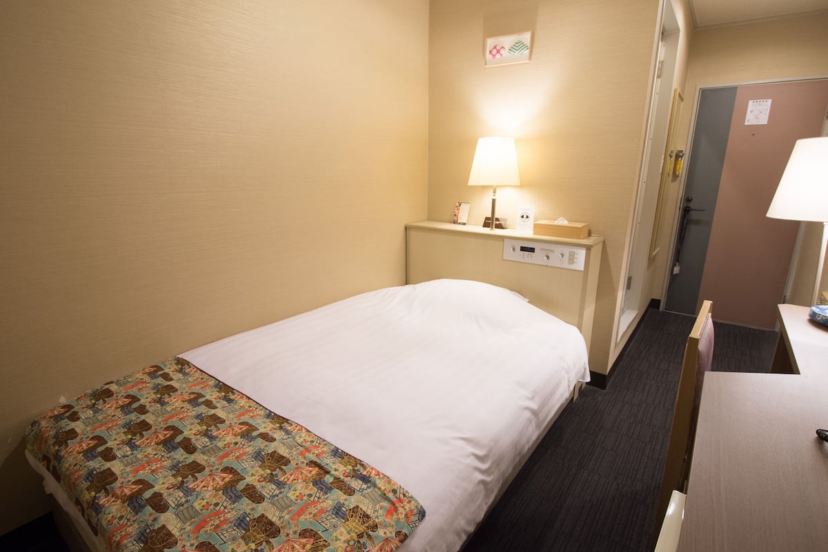 Habitación de hotel con cama, mesita de noche con lámpara, panel de control y puerta.