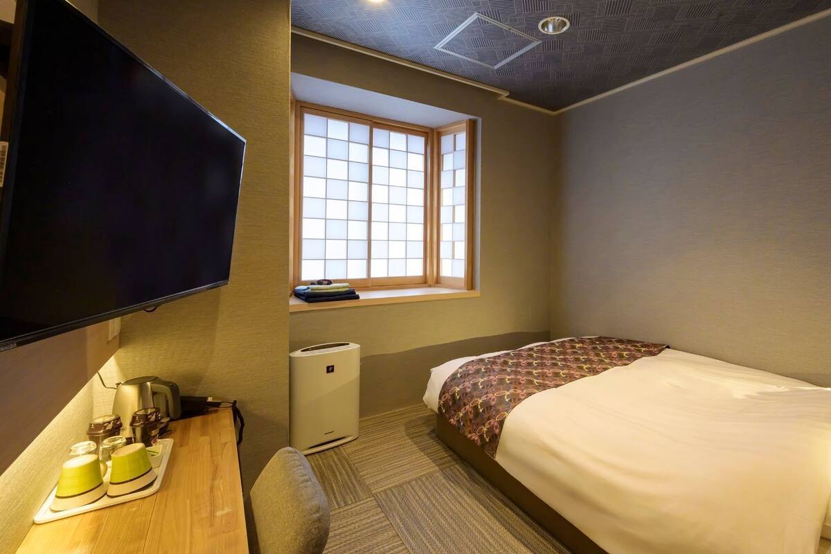 Habitación de hotel con cama, televisión, ventana, escritorio con tetera y un pequeño calentador.