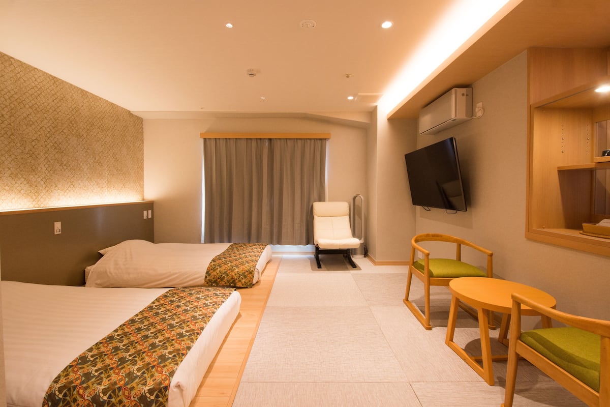 Habitación de hotel con dos camas, una silla, una mesita pequeña, televisión y ventana con cortinas.
