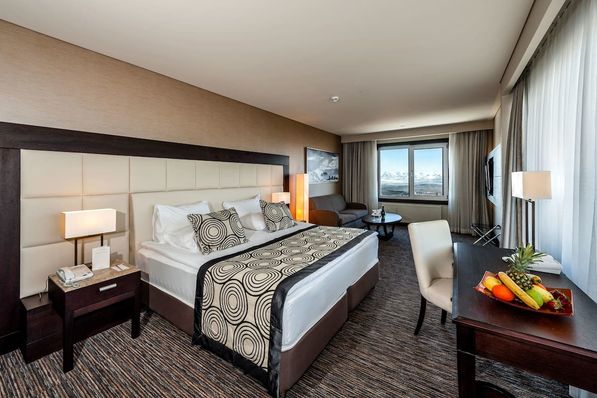 A hotel room with a large bed, a desk with a fruit bowl, a chair, a small table, and a view of the mountains through the window.