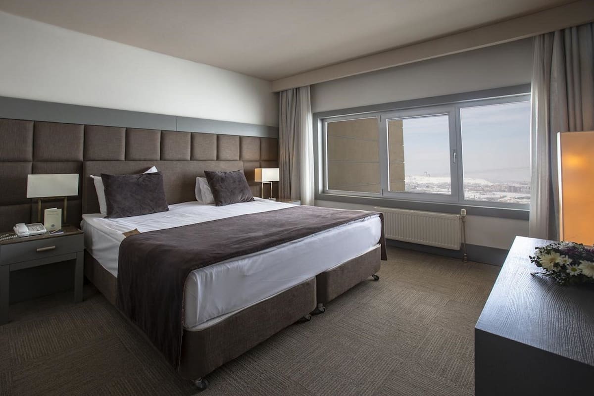 A hotel room with a large bed, two bedside tables with lamps, a view of the city through a large window, and a small table with a vase of flowers.