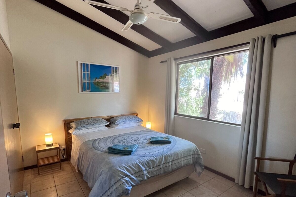 A bedroom with a bed, a ceiling fan, a window with curtains, a nightstand with a lamp, and a painting on the wall.