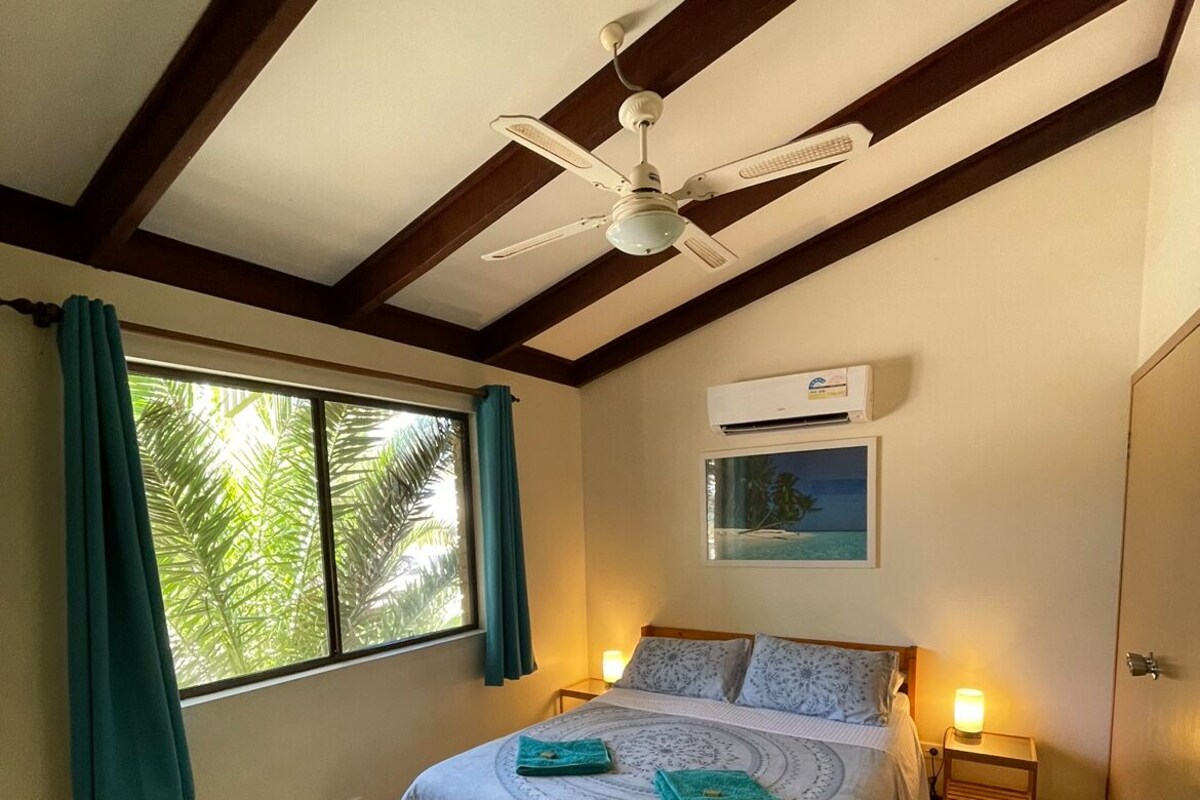 A bedroom with a bed, a ceiling fan, an air conditioning unit, a window with curtains, and a small bedside table with a lamp.