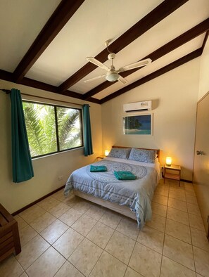 A bedroom with a bed, a ceiling fan, an air conditioning unit, a window with curtains, and a small bedside table with a lamp. - Wildsights Villas (Denham)