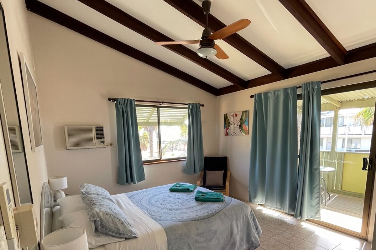 A bedroom with a bed, a ceiling fan, a window with curtains, a chair, and a view of the outside.