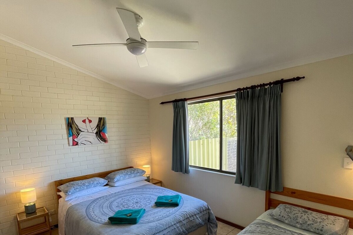 A bedroom with two beds, a ceiling fan, a window with curtains, and a wall-mounted artwork.