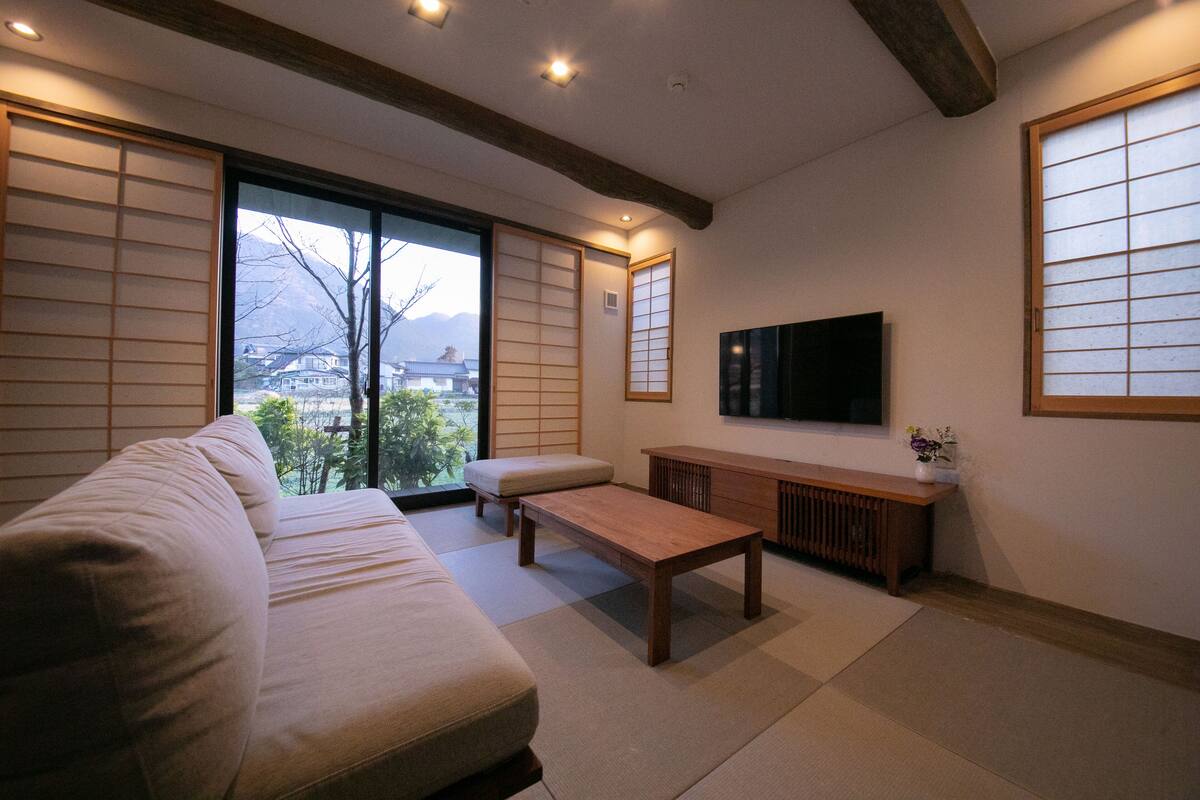 A traditional Japanese-style living room with tatami flooring, a low wooden table, a sofa, sliding shoji doors, and a TV mounted on the wall.