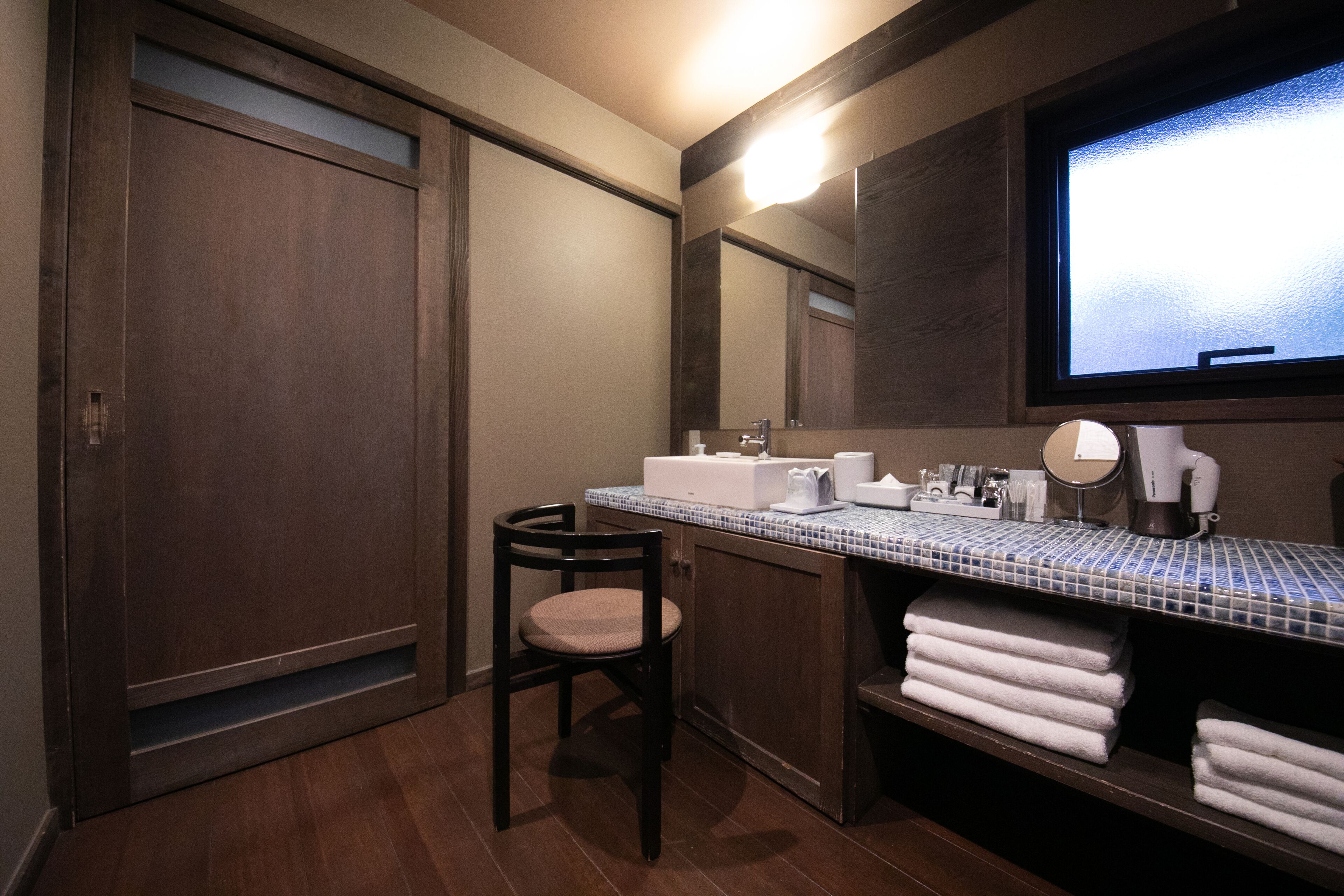 Baño con tocador de madera, espejo, lavamanos y una silla. Hay toallas apiladas a un lado.