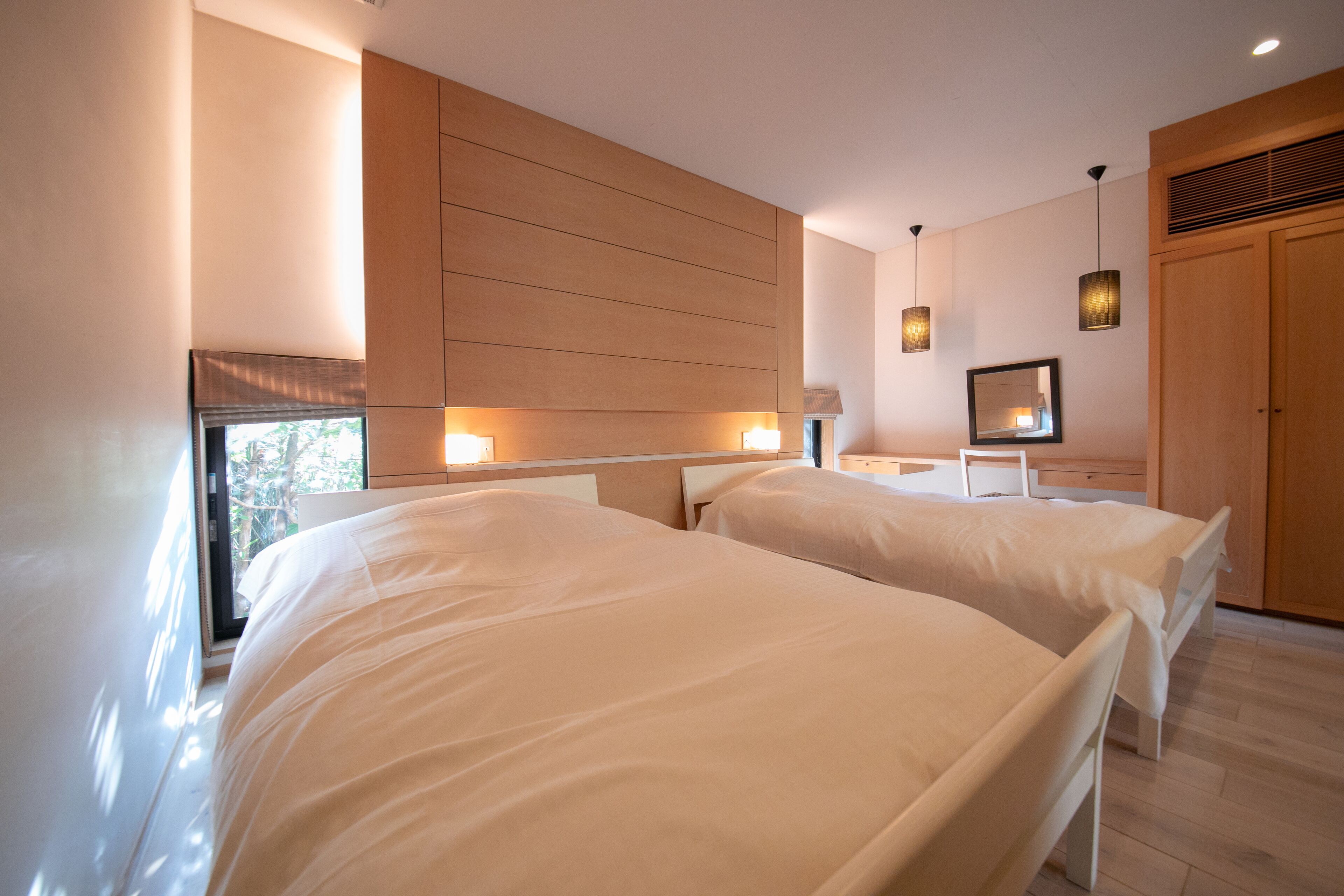 A modern bedroom with two beds, wooden paneling on the wall, pendant lights, and a large mirror.