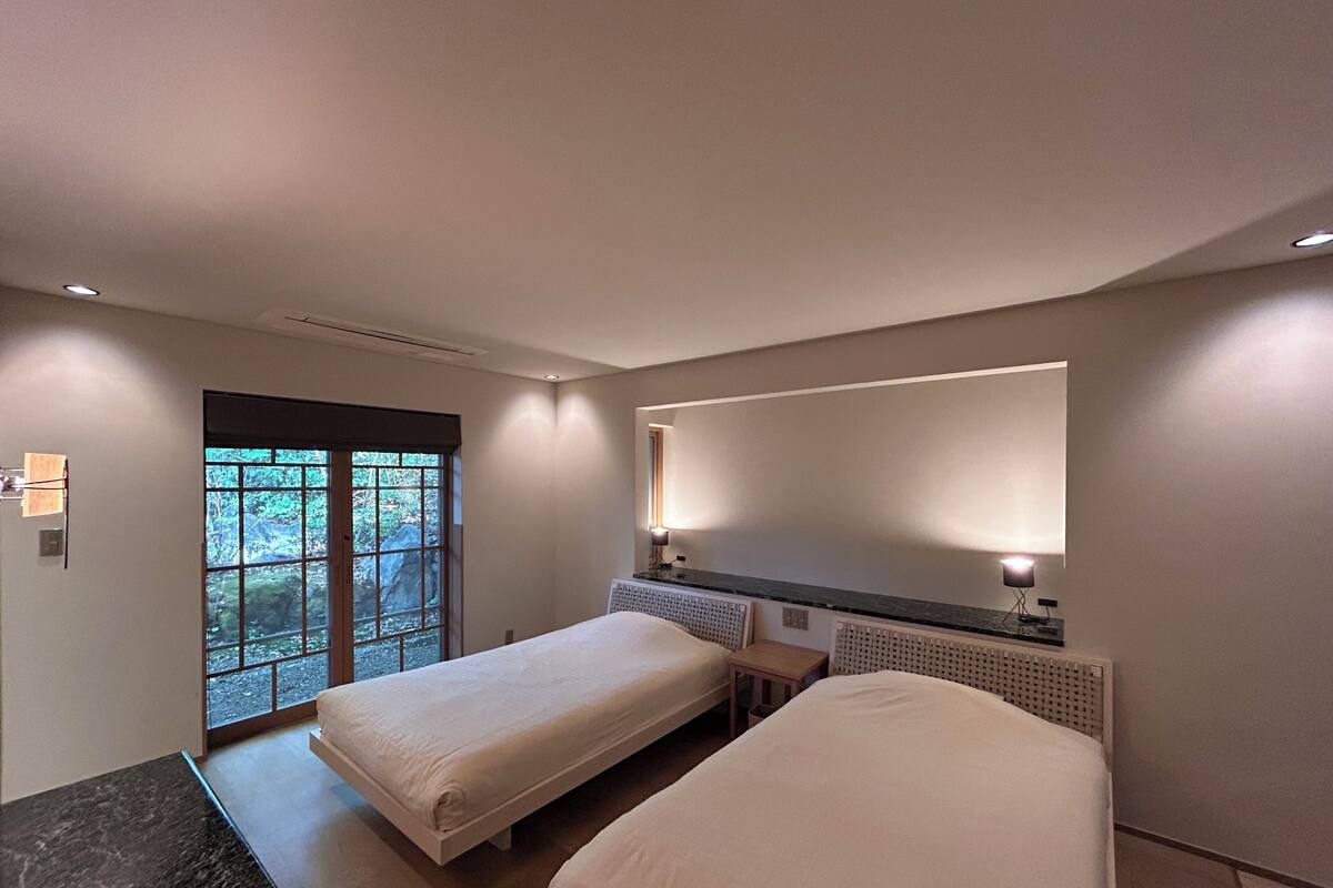 A modern hotel room with two single beds, a small desk, a large mirror, and a view of greenery through the window.