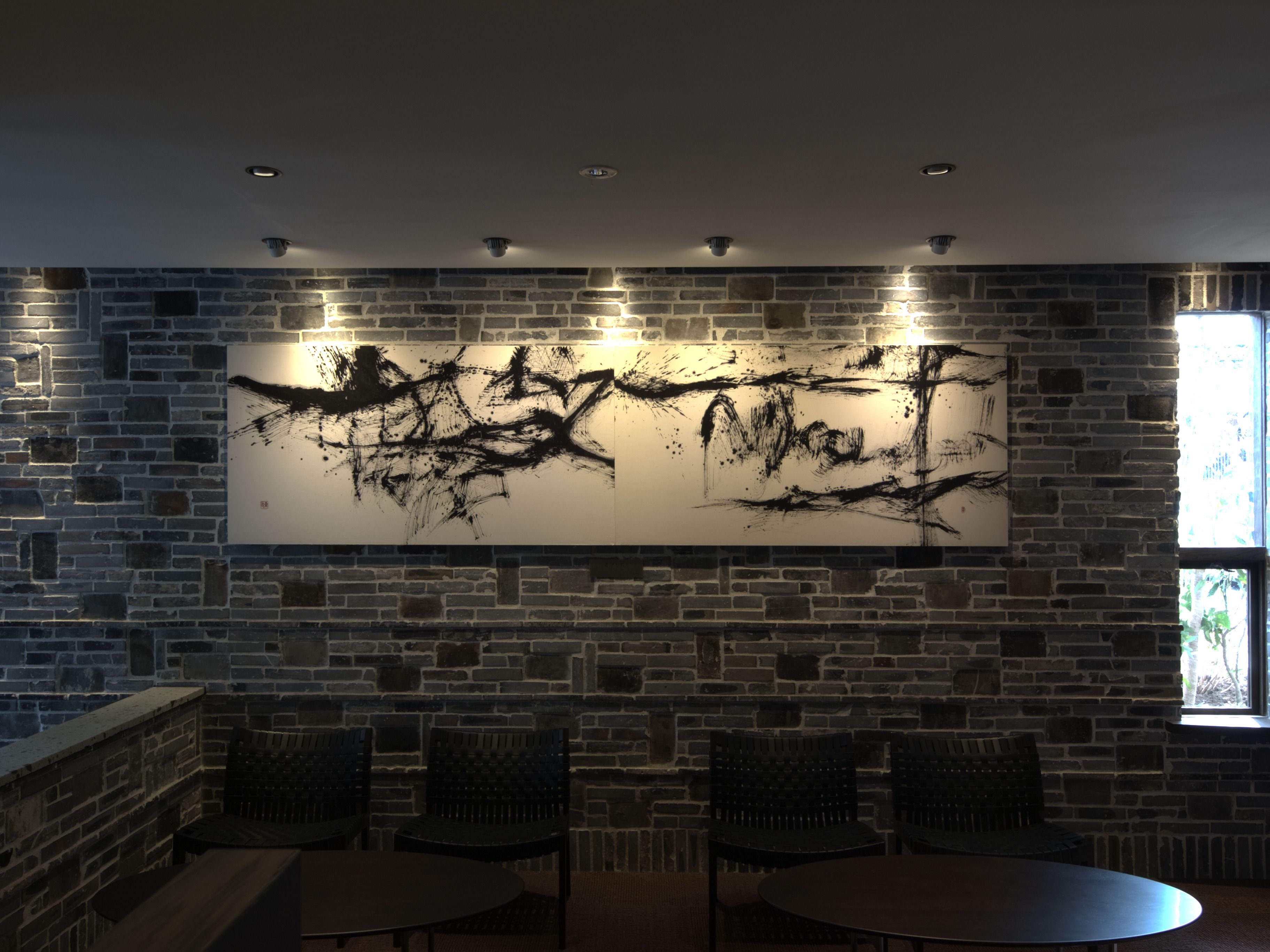 A room with a large abstract painting on a brick wall, a window with natural light, and a seating area with tables and chairs.