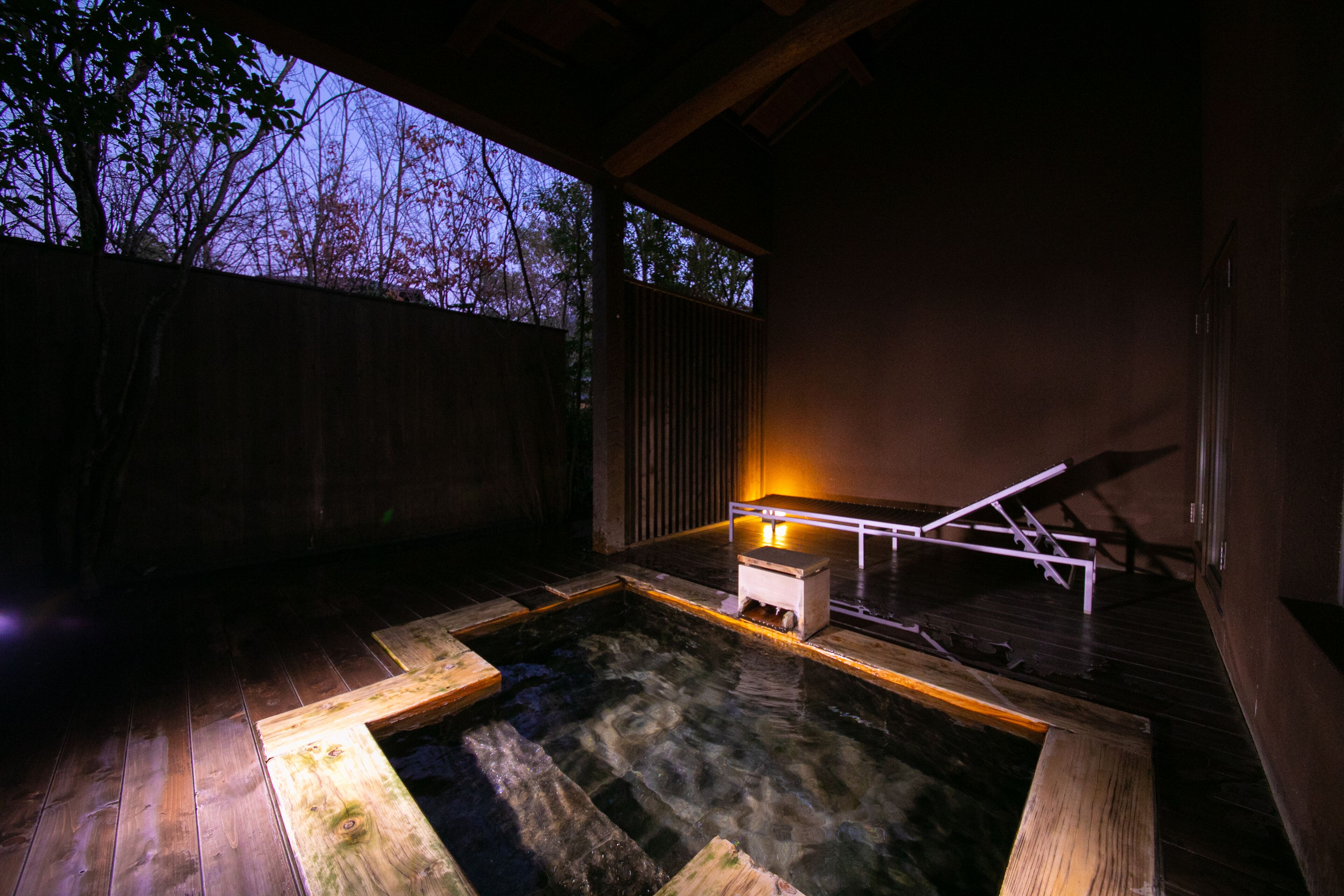A wooden hot tub with a small white structure in the center, surrounded by a wooden deck and a wooden wall.