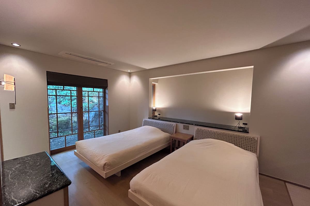 Habitación de hotel con dos camas, una mesita, un escritorio, una ventana con vistas y una lámpara fijada a la pared.