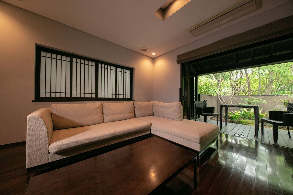A modern living room with a beige sectional sofa, a dark wooden coffee table, and a view of a garden through a large window.