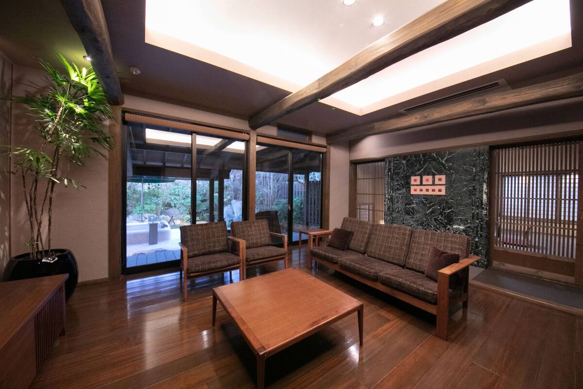 A spacious living room with wooden flooring, a sofa set, and a coffee table. Large windows provide a view of the outdoors.
