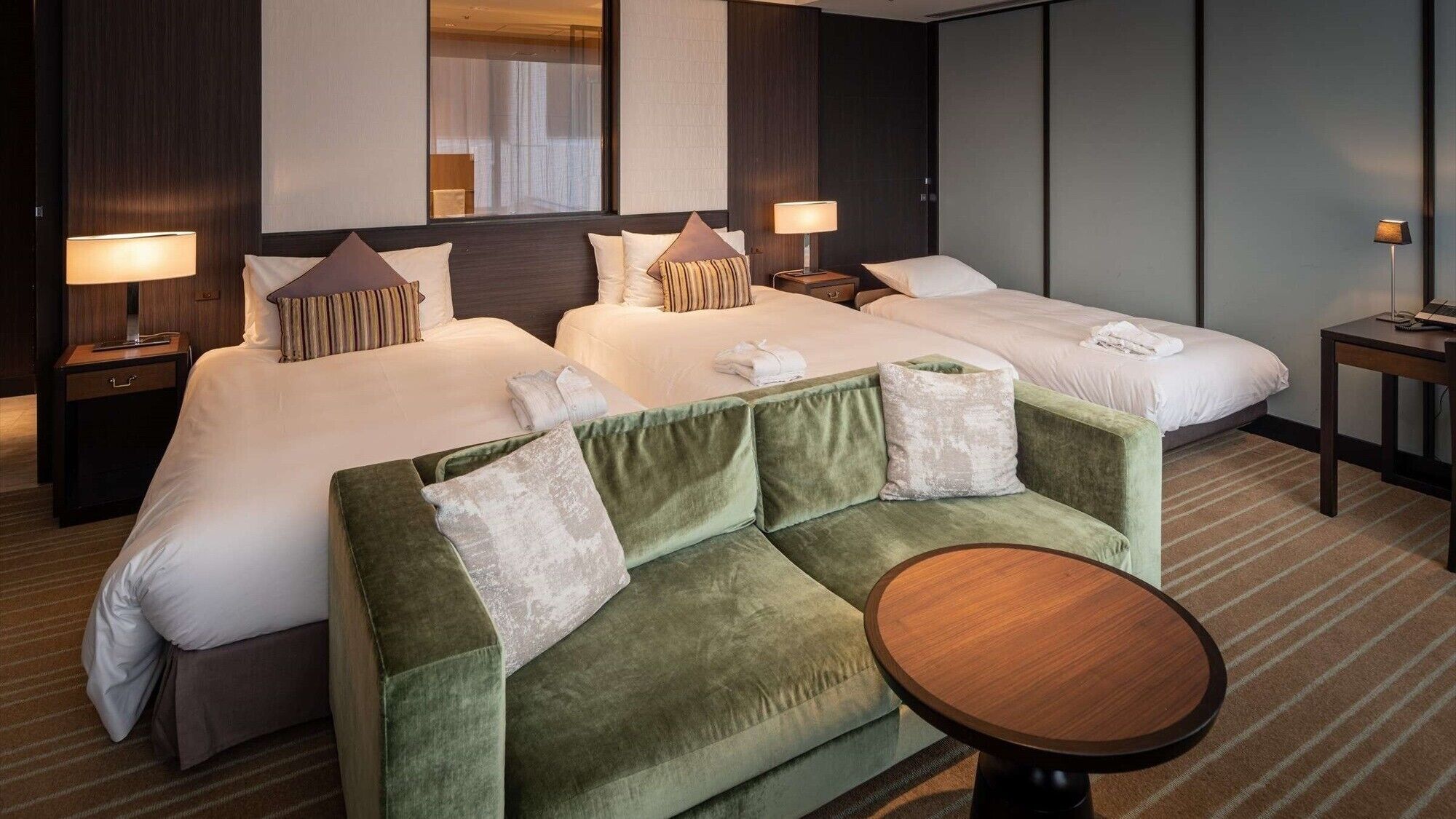 A hotel room with two beds, a green sofa, a small round table, and a nightstand with a lamp.