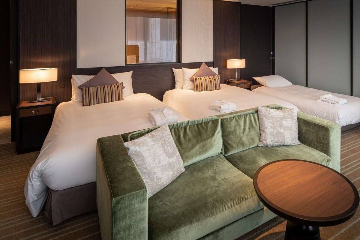 Una camera d'albergo con due letti, un divano verde, un piccolo tavolino rotondo e un comodino con una lampada.