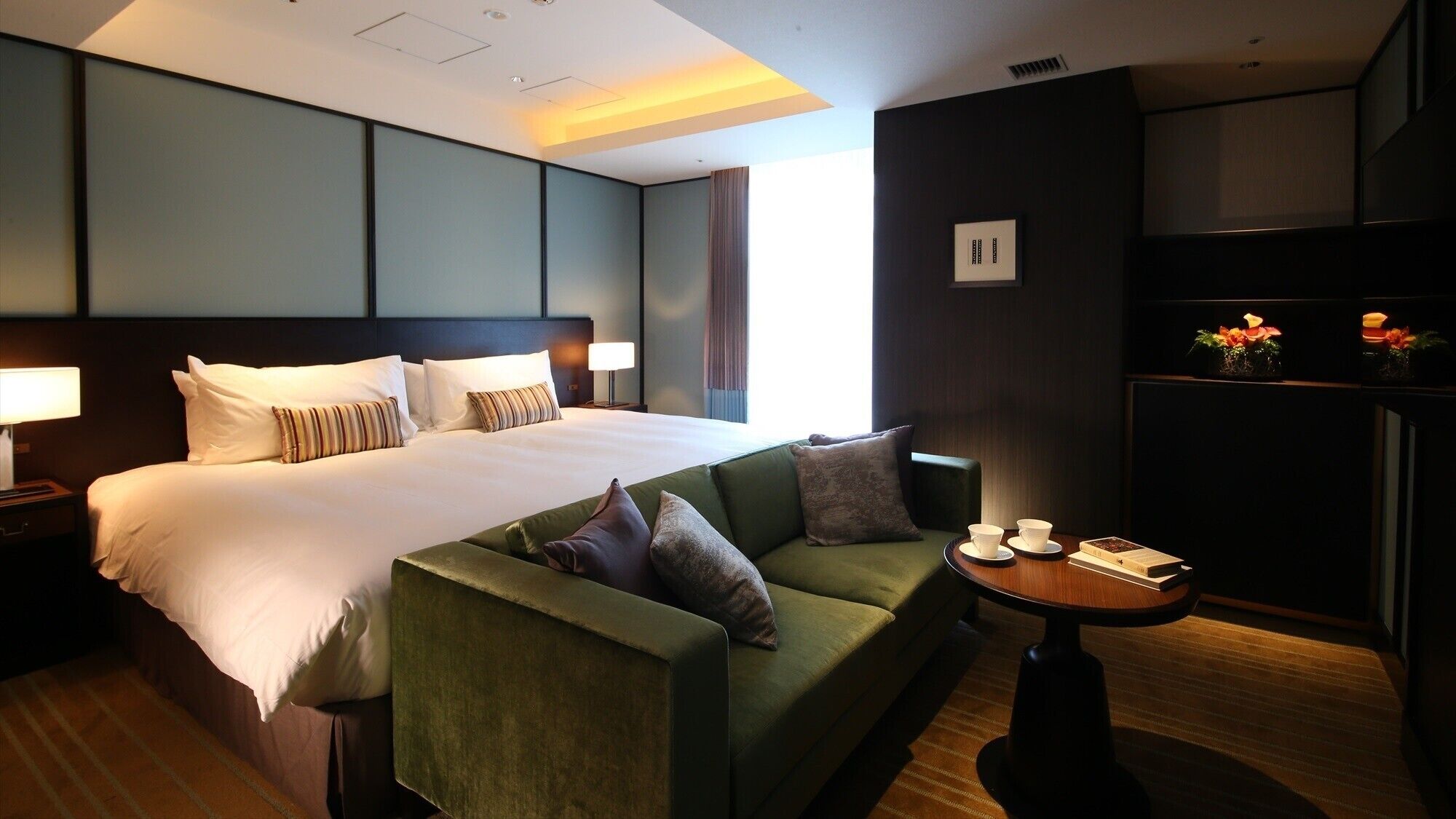 A hotel room with a large bed, a green sofa, a small table with cups and a book, and a wall-mounted TV.