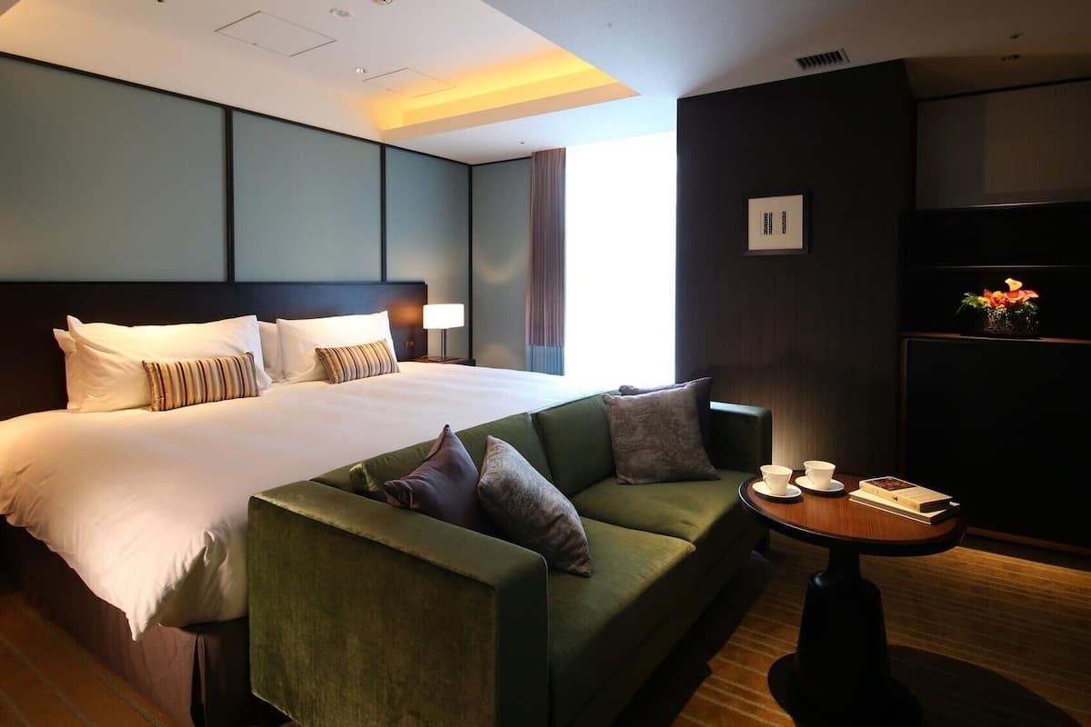 A hotel room with a large bed, a green sofa, a small table with cups and a book, and a wall-mounted TV.
