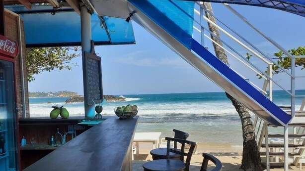 Un bar en la playa con vista al océano, un refrigerador de Coca-Cola y un cartel con la carta.
