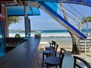 Un bar en la playa con vista al océano, un refrigerador de Coca-Cola y un cartel con la carta.
