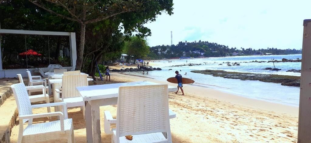 A beachside seating area with white chairs and tables, a person carrying a surfboard, and a view of the ocean and distant hills.
