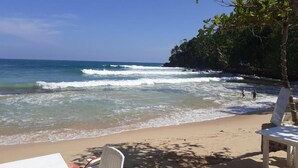 Una playa con sillas de descanso blancas y una mesa, cielo despejado y olas rompiendo sobre la orilla.