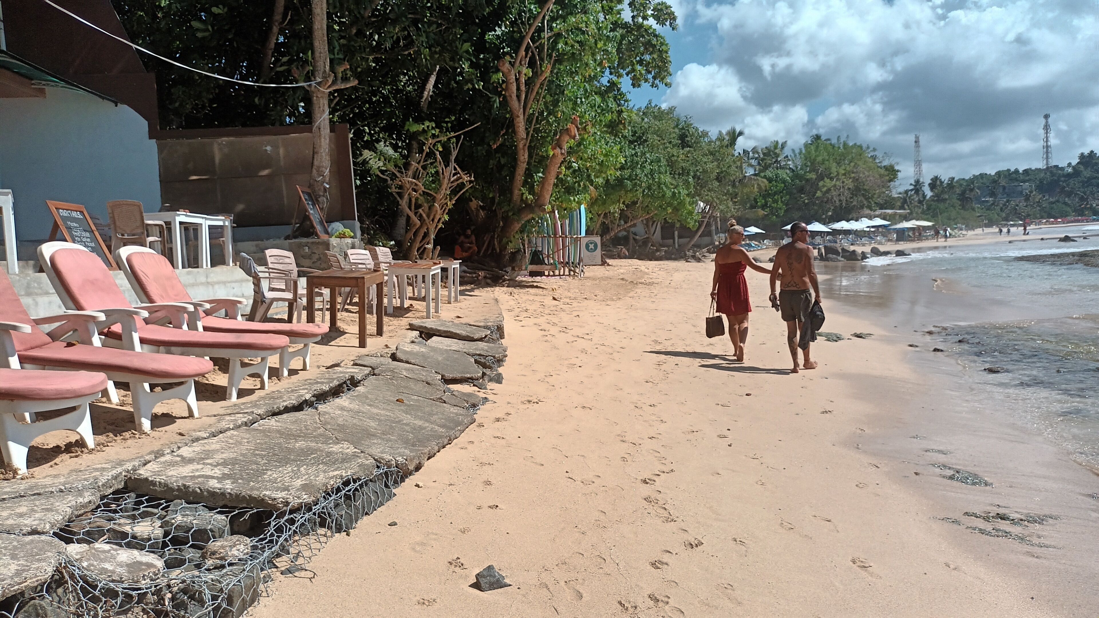 Una zona sulla spiaggia con sedie a sdraio e tavoli, un sentiero sabbioso e vista sull'oceano.